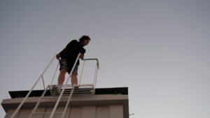 Homeowner steadies a ladder beside a dry roof while inspecting shingles and carrying basic safety equipment.